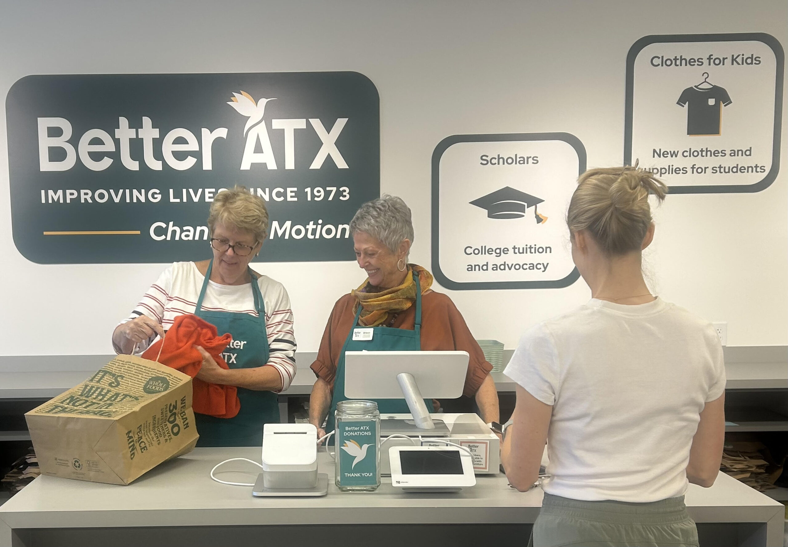 Two women in aprons assist a customer at a checkout counter in the Better ATX clothing donation center.